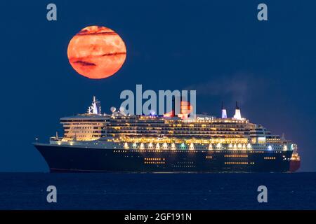 Weymouth, Dorset, Großbritannien. August 2021. Wetter in Großbritannien. Der volle Sturgeon-Mond steigt in den fast klaren Nachthimmel hinter dem Cunard-Kreuzschiff Queen Mary 2, das in der Bucht von Weymouth in Dorset vor Anker liegt. Bildnachweis: Graham Hunt/Alamy Live News Stockfoto