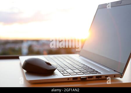 Laptop und kabellose Maus bei Sonnenaufgang auf dem Tisch gegen das Fenster. Gemütlicher Arbeitsplatz im Home Office, Konzept der Arbeit am frühen Morgen Stockfoto