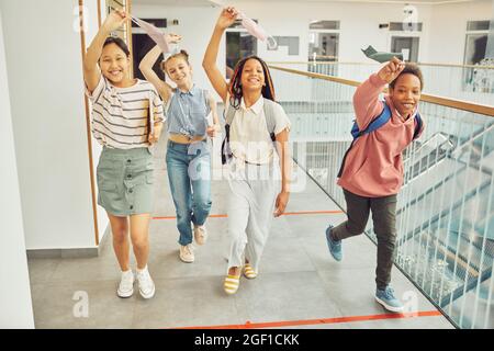 Porträt einer multiethnischen Gruppe von Schulkindern, die Masken halten und fröhlich lächeln, während sie drinnen zur Kamera gehen Stockfoto