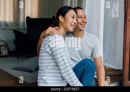 Portrait eines jungen erwachsenen asiatischen Paares, das sich zusammen mit häuslicher Innenausstattung im Hintergrund umarmt. 30s glücklich reifen Mann und Frau lächeln und wegschauen. Ehe und glückliche Beziehung Leben Konzept Stockfoto