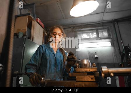 Die junge Frau in ihrer Garage arbeitet an einer Drehbank. Beruf Concept Turner, Metallbearbeitung, Drehen, Industrie, Metall. Stockfoto