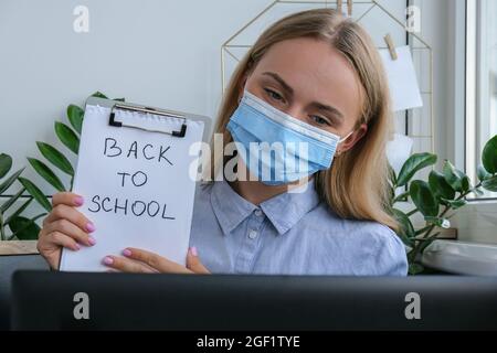 Junge Lehrerin mit medizinischer Gesichtsmaske am Schreibtisch sitzend und mit Laptop zu Hause. E Bildung Zurück in die Schule. Schüler lernen online. Fernunterricht. Prüfungsvorbereitung. Sperren Quarantäne neuer Normalwert Stockfoto