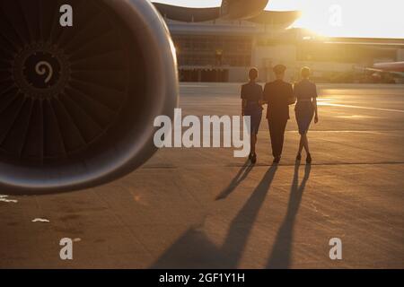 Ganzkörperaufnahme eines professionellen Piloten, der bei Sonnenuntergang zusammen mit zwei weiblichen Flugbegleitern in blauer Uniform im Terminal läuft Stockfoto