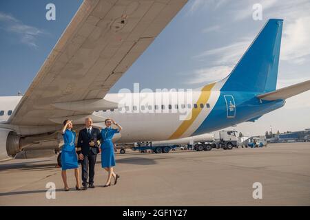 In voller Länge ein glücklicher Pilot, der an einem sonnigen Tag nach der Landung zusammen mit zwei Stewardessen in hellblauer Uniform vor einem Flugzeug läuft Stockfoto