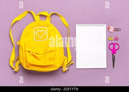 Zurück zur Schule, Bildungskonzept. Gelber Rucksack mit Schulbedarf - Notizbuch, Stifte, Clips, Schere isoliert auf violettem Hintergrund Draufsicht Copy Stockfoto