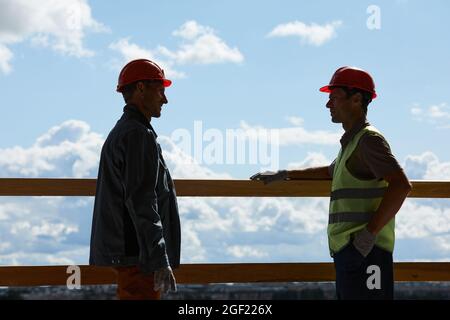 Hintergrundbeleuchtete Seitenansicht von zwei Bauarbeitern, die Hardhuts tragen und sich unterhalten, während sie im Hintergrund gegen den Himmel stehen und Platz kopieren Stockfoto