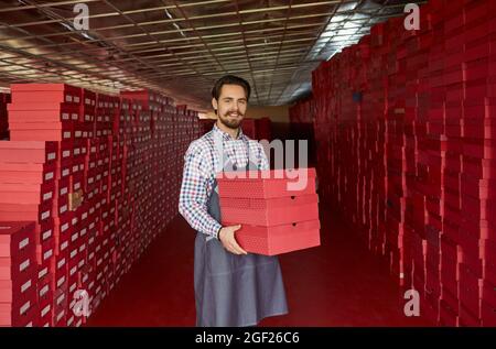 Glücklicher junger Arbeiter, der im Schuhlager steht und Kisten mit neuen Schuhen hält Stockfoto