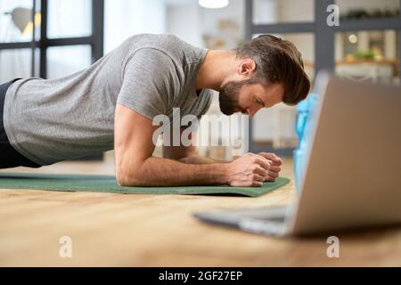 Sportlicher junger Mann, der zu Hause einen Laptop benutzt und eine Planke macht Stockfoto