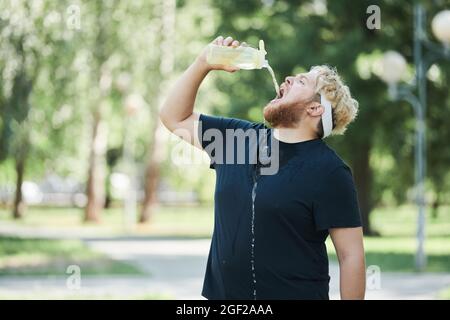 Übergewichtiger Mann, der während seines Sporttrainings im Park Wasser aus der Flasche trinkt Stockfoto