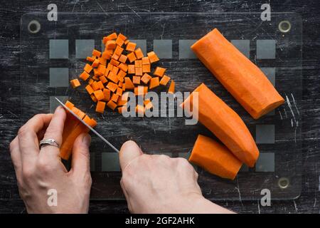 Draufsicht auf weibliche Hände, die rohe Karotte auf kleinen quadratischen Scheiben mit einem Messer auf schwarzem Hintergrund schneiden Stockfoto