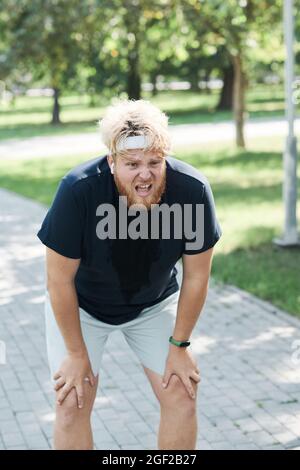 Übergewichtiger junger Mann, der nach dem Laufen im Park atmet und sich ausruht Stockfoto