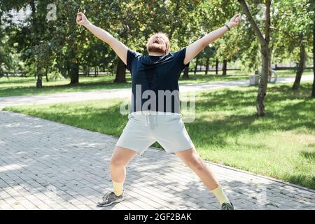 Übergewichtiger Mann beim Training im Freien steht er mit den Armen nach oben und ist zufrieden mit seinen guten Ergebnissen Stockfoto