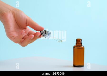 Die weibliche Hand hält das ätherische Öl, das aus dem bernsteinfarbenen Glastropfer fällt. Flasche kosmetisches Öl mit einer Pipette. Speicherplatz kopieren. Hellblauer Hintergrund. Stockfoto