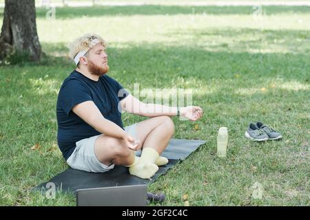 Übergewichtiger Mann, der mit geschlossenen Augen auf einer Trainingsmatte sitzt und im Park Yoga macht Stockfoto