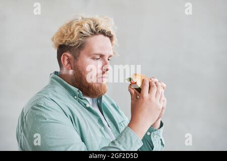 Übergewichtiger Mann, der Burger in den Händen mit traurig vor dem weißen Hintergrund sieht, sitzt er auf einer Diät Stockfoto