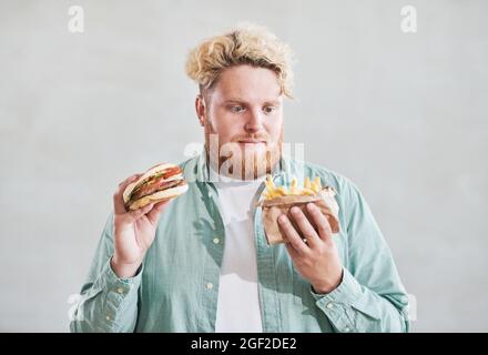 Übergewichtiger junger Mann mit pommes in der einen Hand und Burger in der anderen Hand isoliert auf weißem Hintergrund Stockfoto