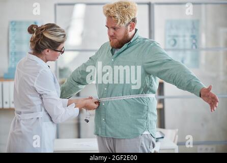 Junge Ernährungswissenschaftlerin, die den übergewichtigen Mann mit einem Zentimeter großen Klebeband misst und eine Diät verschreibt Stockfoto