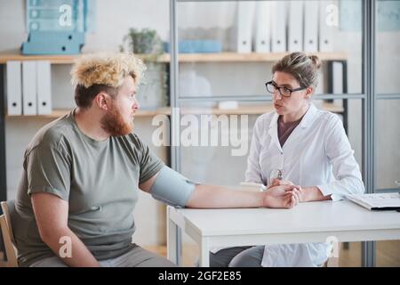 Übergewichtiger Mann, der am Tisch sitzt und mit dem Arzt spricht, während sie während seines Krankenhausaufenthalts seinen Druck misst Stockfoto