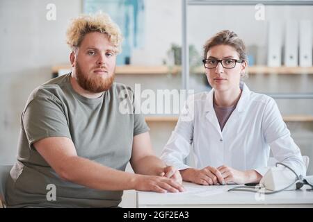 Porträt eines jungen übergewichtigen Mannes, der mit seinem Ernährungsberater am Tisch sitzt und im Büro gemeinsam auf die Kamera schaut Stockfoto