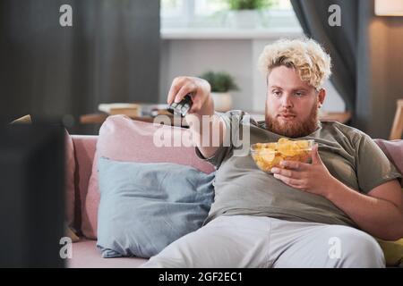 Fauler, übergewichtiger Mann, der mit Chips auf dem Sofa sitzt und die Fernbedienung benutzt, um zu Hause fernzusehen Stockfoto