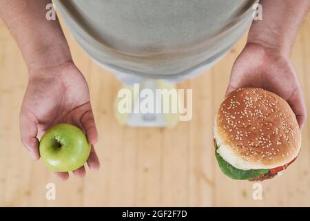 Nahaufnahme eines Mannes, der auf einer Waage steht und apfel in der einen Hand und Burger in der anderen Hand hält, um eine schwierige Wahl zu treffen Stockfoto