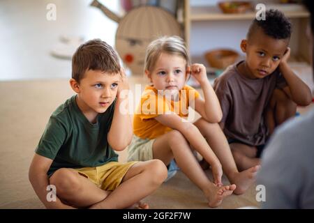 Gruppe von kleinen Kindergartenkindern, die im Klassenzimmer auf dem Boden sitzen und dem Lehrer zuhören. Stockfoto