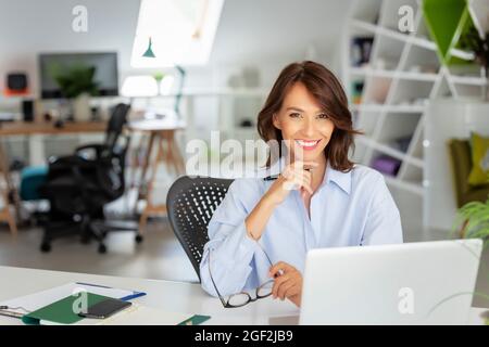 Selbstbewusste Geschäftsfrau mittleren Alters in hellblauem Hemd, sitzt am Schreibtisch hinter ihrem Laptop und arbeitet an einem neuen Projekt. Stockfoto
