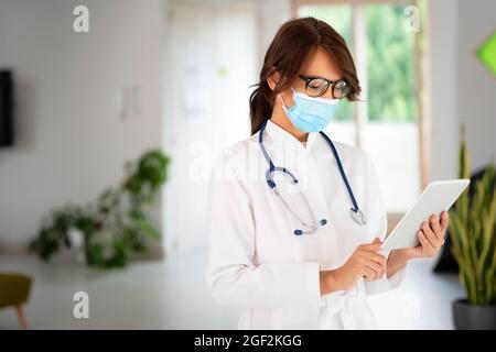 Porträt einer Ärztin, die eine Gesichtsmaske trägt und ihre Patientenakte auf einem digitalen Tablet hält, während sie auf dem Krankenhausflur steht. Stockfoto