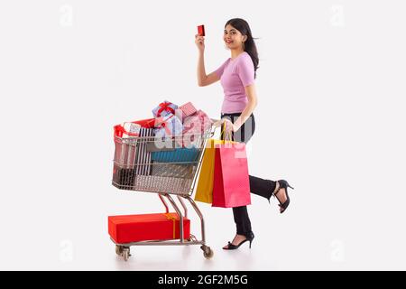 Eine junge Frau mit Kreditkarte und bunten Einkaufstaschen, die einen Wagen voller Geschenkboxen bewegt. Stockfoto