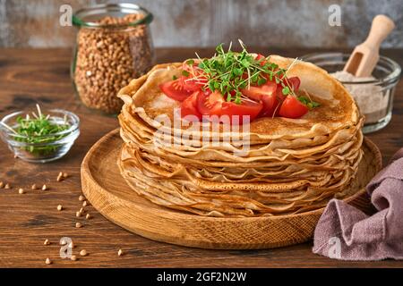 Stapel glutenfreier Buchweizenmehl-Crepes-Pfannkuchen mit Kirschtomaten und Rucola-Microgreens auf Holzplatte, hausgemachtes gesundes Backen zum Frühstück Stockfoto