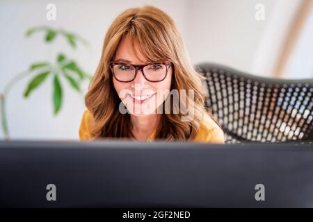 Ausgeschnittene Aufnahme einer Geschäftsfrau, die hinter dem Bildschirm des Computers sitzt, während sie von zu Hause aus arbeitet. Heimbüro. Stockfoto