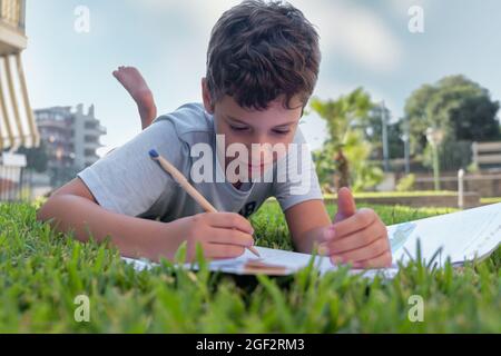 Niedlicher Junge macht Hausaufgaben auf Gras liegen. Kind liest ein Buch im Sommerpark. Konzept der Kinder lernen, studieren, im Freien im Park. Stockfoto