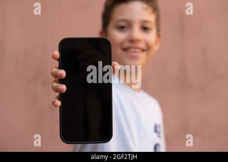 Fröhlicher kleiner Junge mit Smartphone mit leerem schwarzen Display. Verschwommenes Gesicht auf dem Hintergrund isoliert auf einer leeren Wand. Kinder zeigen Mob Stockfoto