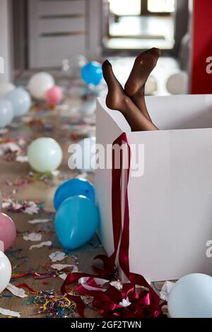 Frau Beine kleben aus großen Box mit undichten Strümpfen, nach Party Chaos, chaotisch im Wohnzimmer zu Hause, Konfetti und Ballons, Morgen nach Party cel Stockfoto