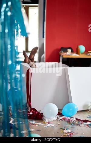 Frau Beine kleben aus großen Box mit undichten Strümpfen, nach Party Chaos, chaotisch im Wohnzimmer zu Hause, Konfetti und Ballons, Morgen nach Party cel Stockfoto