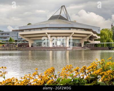 Die brutale Architektur der Central Hall an der York University, eingerahmt von einem See und lebendigen gelben Blumen am Rand, unter einem launischen grauen Himmel. Stockfoto