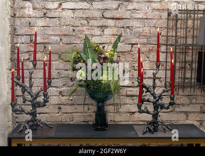 Blumenstrauß in eleganter Vase und zwei Designer-Kerzenständer mit brennenden Kerzen in stilvoller Raumeinrichtung. Loft-Stil Dekorelemente auf Backstein Wand Hintergrund, Loft-Wohnung. Stockfoto