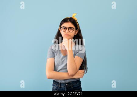 Nachdenkliches indisches Teenager-Mädchen in Brille erinnert sich an etwas und berührt ihr Kinn über blauem Studiohintergrund Stockfoto