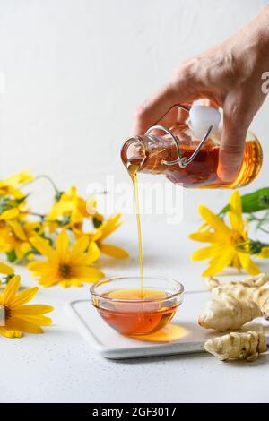 Gießen Jerusalem Artischockensirup in Schüssel, Blumen und Wurzel auf weißem Hintergrund. Vertikales Format. Zuckerersatz für rohe vegane Desserts. Natürliche Swe Stockfoto