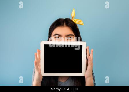 Indisches Teenager-Mädchen versteckt sich hinter Tablet-Computer mit leerem Bildschirm auf blauem Hintergrund, Mockup für Website oder App Stockfoto