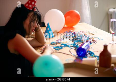 Junge Frau mit rot geriebenem Lippenstift und Kappe, sitzen müde am Tisch in chaotischen Raum nach der Geburtstagsparty, Frau nach der Party zu Hause Stockfoto