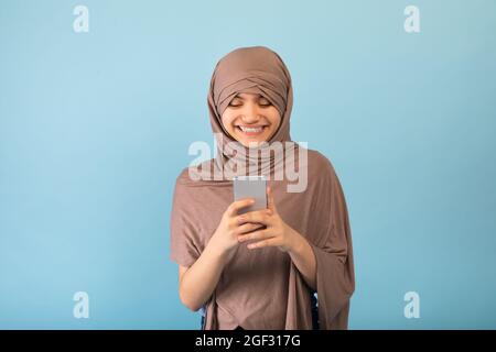 Positive muslimische Teenager-Mädchen in Hijab mit Handy, Nachrichten überprüfen, Surfen in sozialen Medien über blauen Studio-Hintergrund Stockfoto