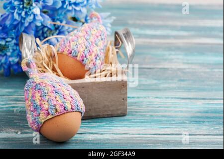Osterkomposition mit Eiern in gestrickten Hüten, blauen Blumen auf einem Holztisch. Stockfoto