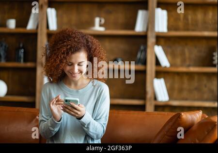 Fröhliche junge rothaarige lockige Frau chattet gerne online, sitzt zu Hause auf der Couch und Messaging auf dem Smartphone. Lächelnde Frau verbringt Freizeit in sozialen Netzwerken, scrollende Feed Stockfoto