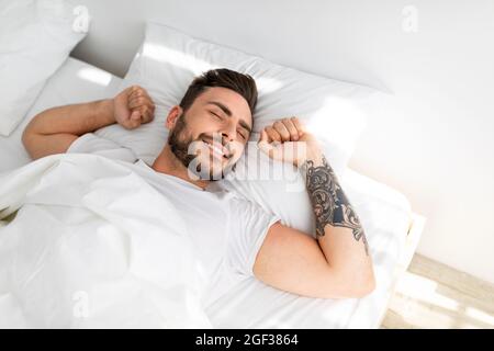 Ein glücklicher, gut gelaunter Kerl, der morgens aufwacht, sich ausdehnt, während er in einem komfortablen Bett liegt und lächelt und einen Blick aus der Höhe hat Stockfoto