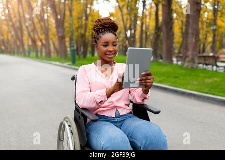 Porträt einer behinderten jungen schwarzen Frau im Rollstuhl, die einen Tablet-pc für die Fernkommunikation im Herbstpark verwendet Stockfoto