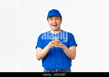Technologie, Online-Shopping und Versandkonzept. Portrait von amüsiert und glücklich asiatischen jungen Delivery Mann mit Zahnspangen, trägt blaue Uniform, suchen Stockfoto