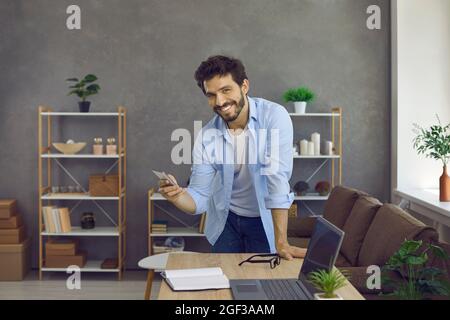 Selbstbewusster junger und fröhlicher Mann, der in seinem Heimbüro mit einem Mobiltelefon in den Händen steht. Stockfoto