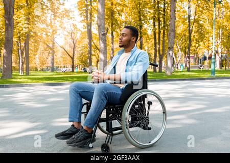 Die ganze Länge des friedlichen schwarzen behinderten Kerl im Rollstuhl, der im Herbst im Stadtpark spazieren gehen musste Stockfoto