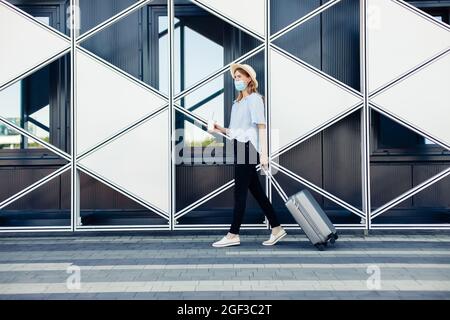 Reisende Tourist Frau in Sommer Freizeitkleidung, trägt medizinische Schutzmaske gegen Corona-Virus, Frau mit Koffer auf dem Hintergrund von schwarz Stockfoto